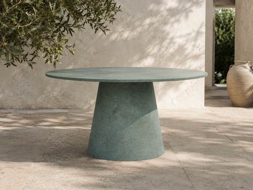 Tavolo ORBIS MANOFATTI in finitura verde salvia ambientato su una terrazza mediterranea in pietra con ombre di ulivo